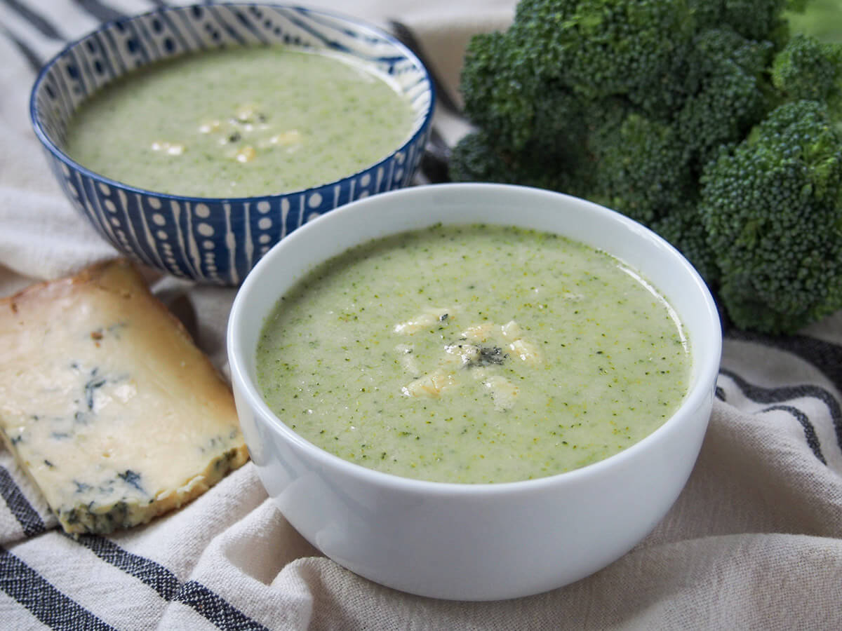 side view of two bowls of broccoli and stilton soup with stilton wedge to side and broccoli behind.