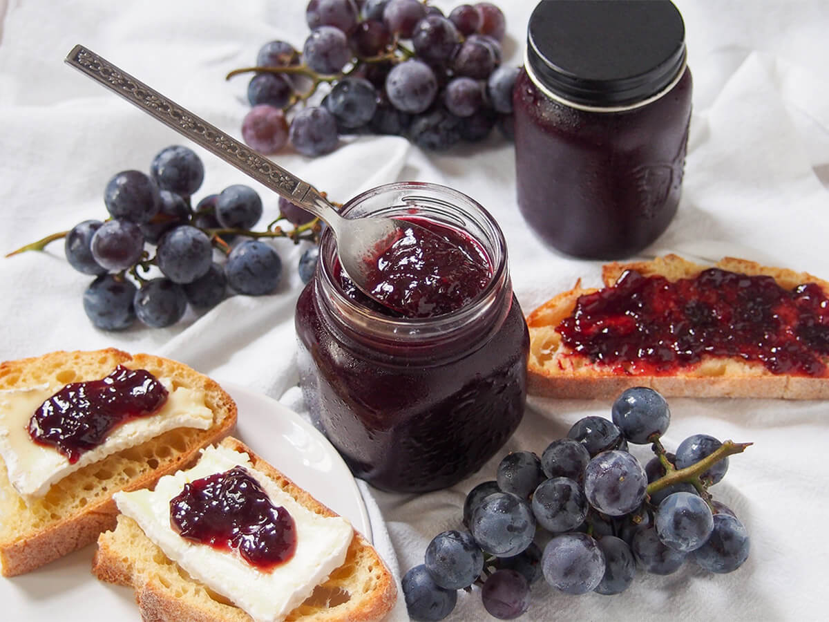 jar of Concord grape jam with piece of bread topped with jam to one side, and bread with cheese and jam on other