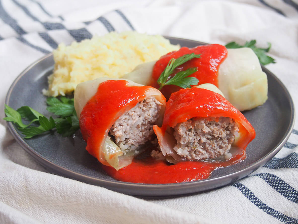 a cut golabki in front of a whole stuffed cabbage roll and mashed potato, sauce over top and parsley garnish.