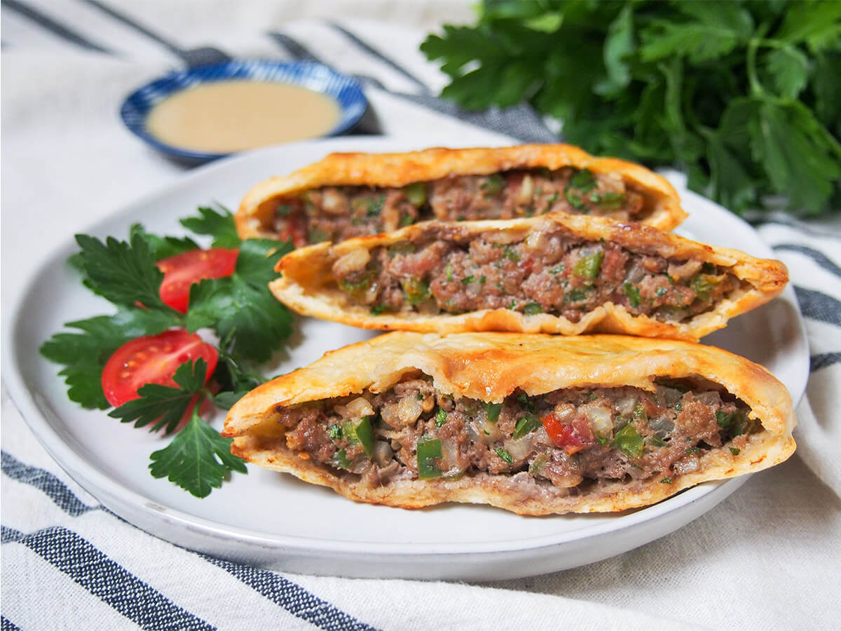 three hawawshi meat filled pita on plate with tomato and parsley to side