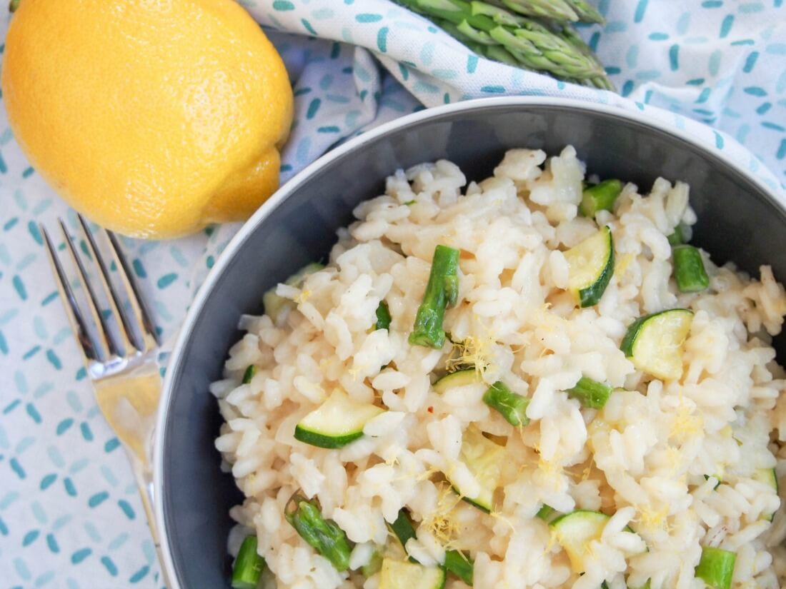 part view of bowl of lemon risotto with asparagus and zucchini with fork to side and lemon beside bowl