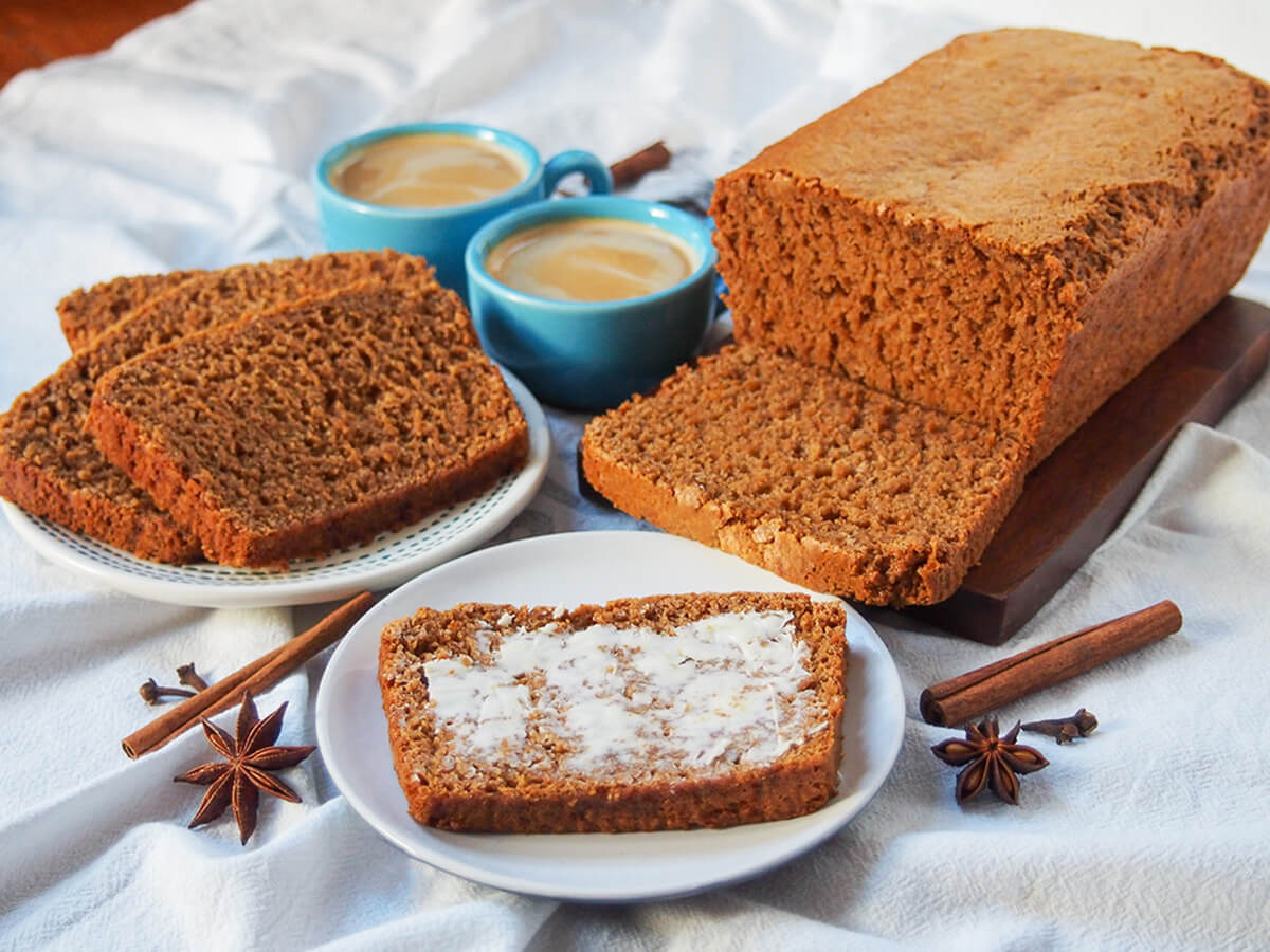 buttered slice of ontbijtkoek Dutch spice cake with loaf and more slices behind and coffee cups in middle back.