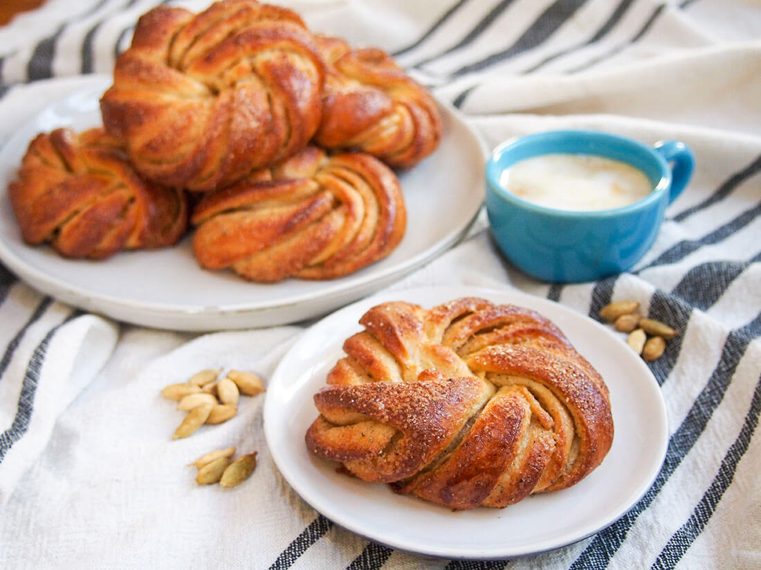 Swedish cardamom buns - one on small plate in front, more on large plate behind with cup of coffee